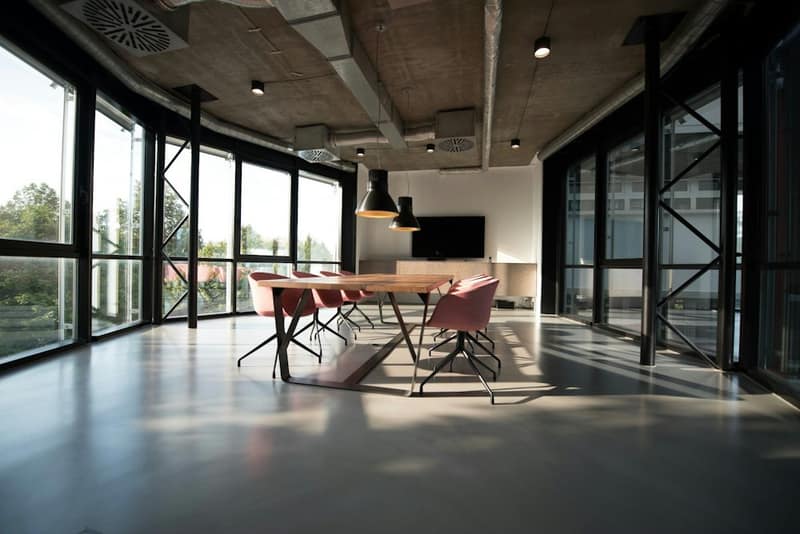 An empty glass-walled meeting room with a long table and pink chairs